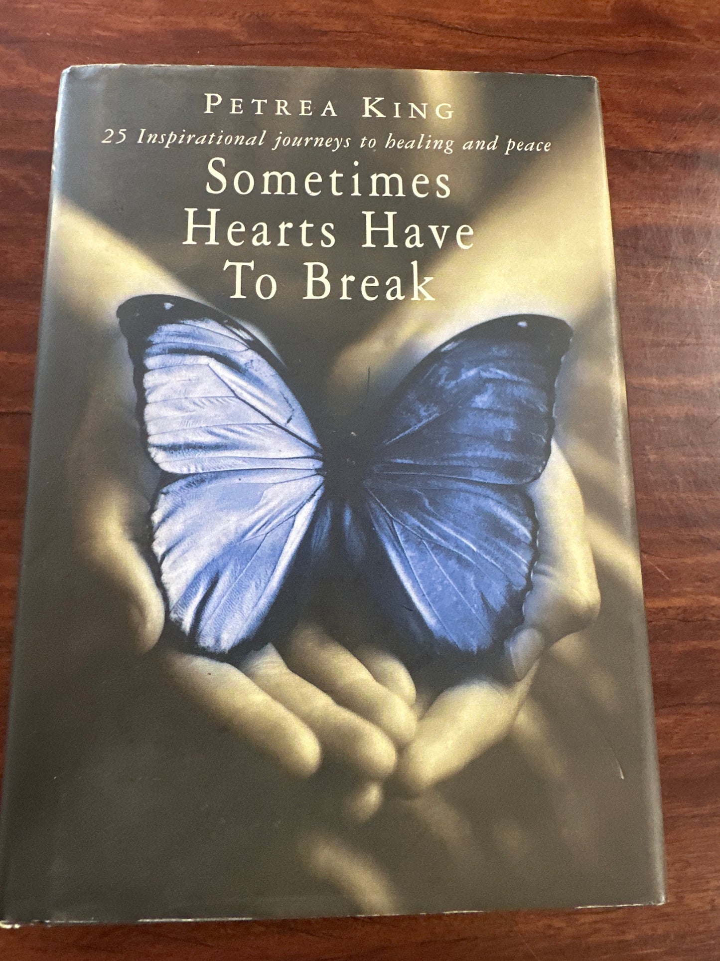 Book cover of Sometimes Hearts Have to Break with a blue butterfly on hands against a wooden background